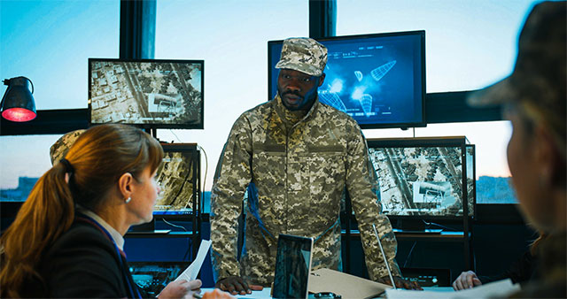 A man in a meeting surrounded by computers displaying military images